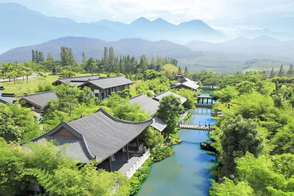 士毛月富贵山庄鸟瞰照片：山脚下的黑瓦中式建筑、河流与绿地，远处是群山